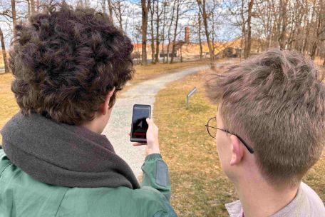 Zwei junge Schüler stehen in der Gedenkstätte am Rand des ehemaligen Zoos. Sie schauen auf ein Handy, das einer der beiden hochhält. Auf dem Handy ist ein Navigationspfeil zu erkennen, der ihnen die Richtung weist. Im Hintergrund sieht man den Lagerzaun und den Schornstein des Krematoriums.