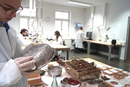 Eine Person reinigt eine alte Essschüssel. Auf dem Tisch vor der Person sind historische Objekte zu sehen. Menschen im Laborkittel im Hintergrund an Geräten.