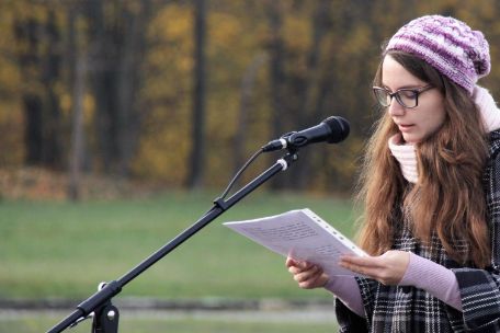 A young woman is standing on a meadow in front of a microphone and is reading from a piece of paper.