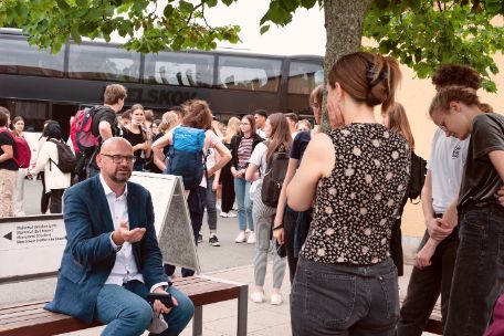 A group at the beginning of a guided tour, listening to a seated memorial staff member. In the background, a school class gets off a bus and gathers in front of the visitor information desk.