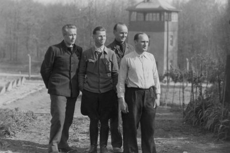 Group photo of four men including Otto Leischnig. In the background you can see a Buchenwald watchtower.