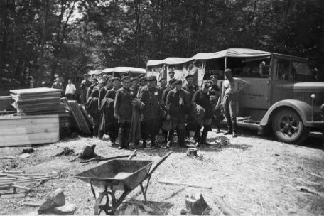 Prisoners lined up in rows of four stand amid tall trees. Next to them lie building materials and a wheelbarrow. In the background, SS guards stand in front of their personnel carriers.