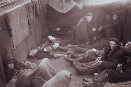 In an open freight car, a dozen starving prisoners lie and cower. Between them lies a deceased man.