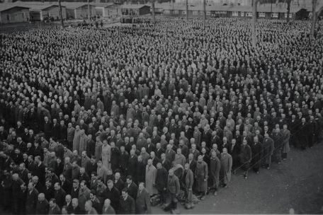 On the crowded Buchenwald roll call square, about a thousand men stand separately in groups of eight. They stand in rows in blocks of about five by 16, but between the blocks there are other people. In the background some barracks.