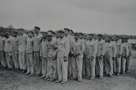 Die in Reihen stehenden Häftlinge tragen alle die gleiche zebragestreifte Häftlingskleidung und Mütze. Einige zeigen eine gebeugte Haltung oder einen schmerzverzerrten Gesichtsausdruck.