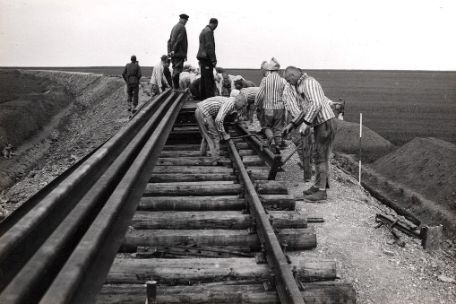 Häftlinge in gestreifter Kleidung beim Verlegen von Gleisen auf einem aus Erde aufgeschichtetem Damm mit einfachem Werkzeug. Im Hintergrund beaufsichtigen SS Mitglieder den Bau.