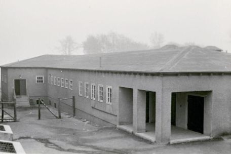 The disinfection building from the outside. A simple, flat building. Two entrances can be seen. A smaller one on the left behind a fence, one on the right in front.
