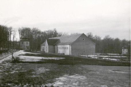 Gebäude der Häftlingskantine. Im Hintergrund links und rechts die Wachtürme Nr. 2 und 3. Dazwischen verläuft der Lagerzaun. Auch das Gelände davor ist von kleineren Zäunen unterbrochen. 