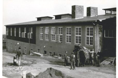 The carpentry building stretches from the front right to the back left. In front of the building runs a dirt road on which, on the right, a group of prisoners is unloading a Karen with boards. To the left, a prisoner carries a bucket to the building.