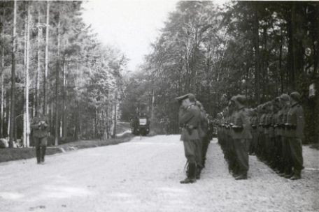 Rechts stehen drei Reihen SS-Männer in Reih und Glied auf einer Fläche aus Kies. Links an der Bahnsteigkante steht ein Offizier. Hinten Links kann man einen einfahrenden Zug erkennen.