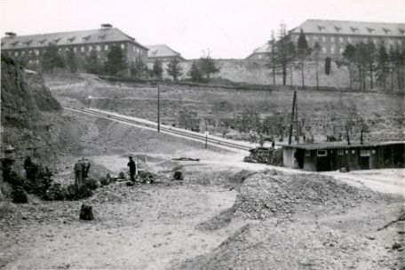 View of the quarry. It runs terraced ascending. In the foreground a small hut. In the background: Waffen SS barracks above the quarry. In the center: Ramp with lorry tracks. To the right and left: prisoners at work.