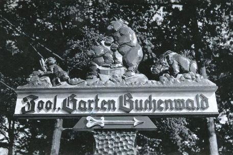 Ein handgeschnitzter Wegweiser mit der Aufschrift "Zool. Garten Buchenwald“, mit Tierfiguren oberhalb des Schriftzuges: Ein Affe, zwei Bären und ein Wildschwein