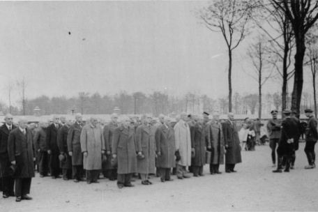 A group of Jewish prisoners in civilian clothes stands in rows of 7 on the roll call square. All heads are shaved. On the right in the picture SS men. In the background, prisoners of the barber work command shave the heads of the newly admitted prisoners.