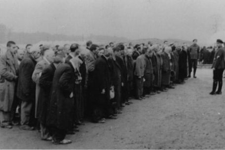 Eine Gruppe von Häftlingen in Zivilkleidung steht auf dem Appellplatz. Vor ihnen ein Uniformierter. Foto von vorne Links.