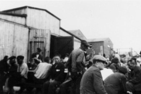 Häftlinge des Kleinen Lagers sitzen und stehen dicht an dicht vor dem offenen Tor einer Pferdestallbaracke. Dabei handelt es sich um Block 51. Dahinter sind die Steingebäude der Blöcke 46 und 41 zu erkennen.