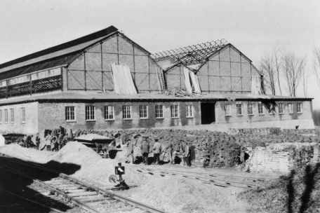 Gleisanschluss der Buchenwald Bahn in den Gustloff-Werken II. Im Hintergrund arbeitende Häftlinge. Vom Werk ist das im Bau befindliche, zweigeteilte Dach einer großen Halle zu sehen. Baumaterial ist an verschiedenen Stellen aufgeschichtet.