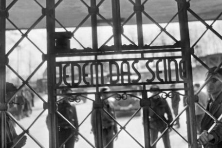 Blick durch das Lagertor auf die Inschrift "Jedem das Seine". Hinter dem Tor amerikanische Soldaten und ehemalige Häftlinge.