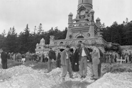 Beisetzung von 1.286 Urnen verstorbener Häftlinge des Konzentrationslagers Buchenwald. Im Hintergrund ist der Bismarkturm zu sehen. Im Vordergrund stehen drei Männer um ein frisch ausgehobenes Grab. Weiter hinten am Bismarkturm eine Menschenmenge.