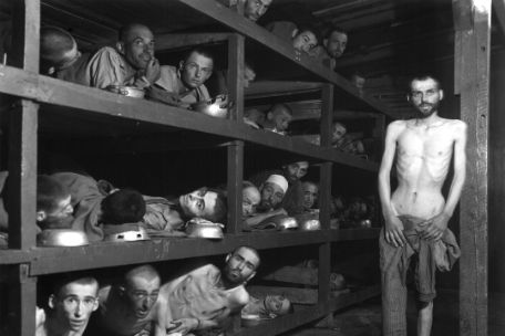 Simon Toncman standing in front on the right, undressed. He holds a jacket in front of the lower half of his body. To the left, other prisoners are lying in shelf-like scaffolding. All are looking into the camera. The men are, as far as can be seen, completely emaciated. Some are wrapped in thin blankets and use metal bowls as headrests.