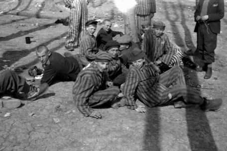 Zehn minderjährige Häftlinge in Sträflingskleidung sitzen auf dem Boden. Einige blicken direkt in die Kamera. Hinter ihnen sind die Beine dreier erwachsener Häftlinge zu sehen, die hinter den Kindern stehen. Der Boden besteht aus trockener, brüchiger Erde und Geröll. 