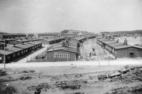 View from watchtower 3 over the rows of barracks of Buchenwald Concentration Camp from west to east. In the foreground, Block 1 can be seen on the right, Block 7 below it, and Block 13 of the Soviet POW camp on the far left. People are walking between the barracks. Broken furniture and other debris are piled up at the front edge of the picture. 