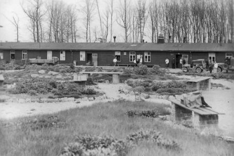The picture shows the garden of the prisoner infirmary with flower beds, benches and branched paths. In the background, the long low building of the prisoners' hospital can be seen. In front of it are isolated nursing staff with white jackets.