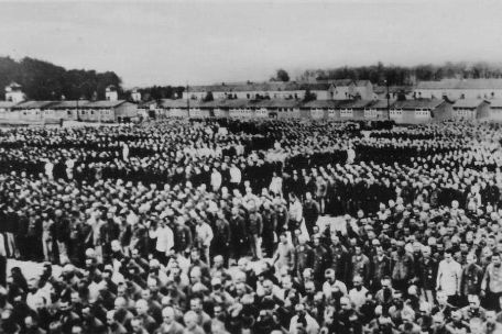 Prisoners of Buchenwald concentration camp lined up in blocks for roll call. There are thousands of them. Individual faces are barely recognizable In the background, the camp barracks. 