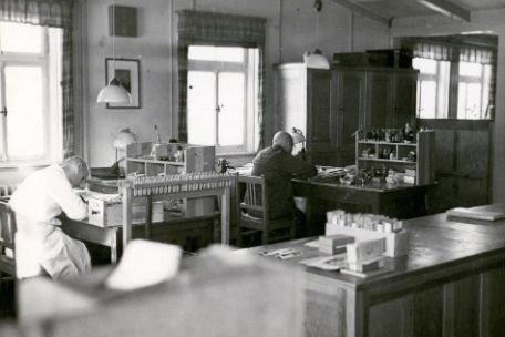 Two prisoners at archival work in the office of the recognition service photographed diagonally from behind. On the right, presumably the prisoner Karl Siebeneichler dressed in white. On the left, another prisoner dressed in black.