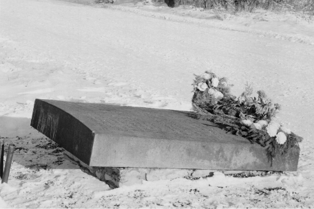 A massive, regularly worked memorial stone from the side. It consists of a large, flat cuboid bearing the inscription, angled at a slight angle. Due to the perspective, the text is not legible. On the right, on the lower end of the cuboid, there is a floral arangmant.