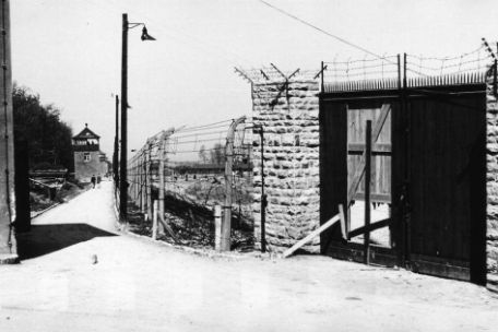On the left, the post path runs straight along the camp fence. Guard towers can be seen at regular intervals along the path. On the right side of the picture, a gate in the camp fence, provisionally closed with wooden boards, can be seen. The fence consists of high posts that are bent inward at the top, into the camp. Barbed wires are stretched between them. 