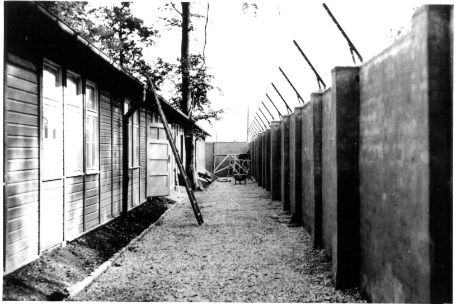 Path between the wall and the isolation hut. Both are about the same height. A ladder leads to the roof of the barrack. The wall is covered with barbed wire. The path ends at another wall.