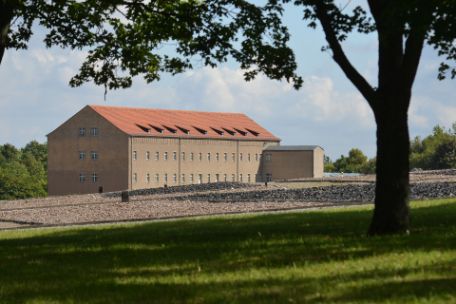 View across the site to the former depot building. It is large and long. In the foreground, the ground plans of the former barracks can be seen in darkness. 