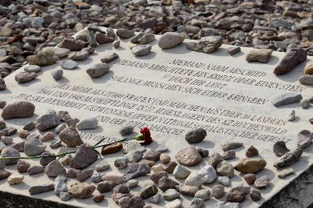 Memorial stone covered with small stones with engraved inscription: "Nobody said goodbye. No one erected a cross or a stone. But you will live as long as people remember you. To the more than 27,000 women and girls from over 30 countries who were forced into slave labor as prisoners in 27 subcamps of Buchenwald concentration camp in 1944 and 1945, victims of Nazi violence and the death marches. Non Omnis Moriar." Under the lettering a single red flower