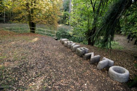 A row of concrete letters that form the word Memento. In the background a balustrade marks an earthen funnel surrounded by trees