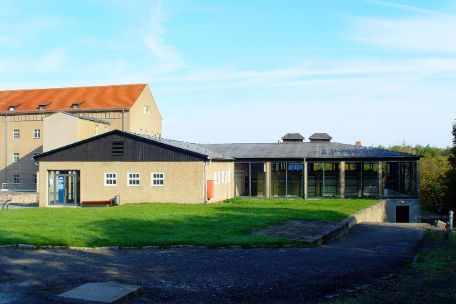 Das flache ehemalige Desinfektionsgebäude in seiner heutigen Gestalt. Der rechte Flügel hat Glaswände. Im Hintergund das ehemalige Kammergebäude.