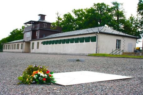 The silver square memorial plate placed on the ground. In front of the plate is a flower arrangement. In the background you can see the gate building.