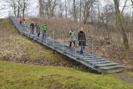 Besucher:innen begehen die Zeitschneise vom Lager aus. Eine Treppe führt zum ehemaligen Postenweg hinab.