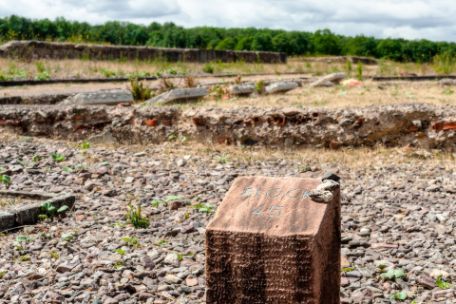 Ein orangefarbener Steinklotz mit der Gravur: "Block 45". Im Hintergrund sind nebeneinander fünf Gedenksteine von hinten, die am ehemaligen Standort des Blocks aufgestellt wurden.