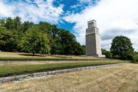 Am leicht abfallenden Hang des oberen Ettersbergs stehen vereinzelte Bäume zwischen mehreren Reihen von Blumenbeeten. Hinter ihnen der Glockenturm der Gedenkstätte.