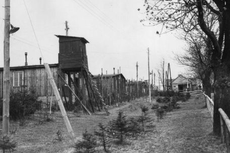 Blick auf einen Wachturm und den Lagerzaun des Buchenwalder Außenlagers Ohrdruf. Der Wachturm wirkt provisorisch und erinnert an einen Jägerstand.