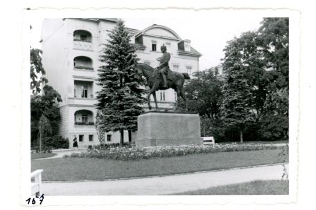 Zu sehen ist ein Denkmal. Auf einem Sockel steht ein Bronzepferd mit Reiter. Um den Sockel des denkmals ist ein Beet angelegt. Im Vordergrung verlauft ein Weg einmal durch den Bildausschnitt.