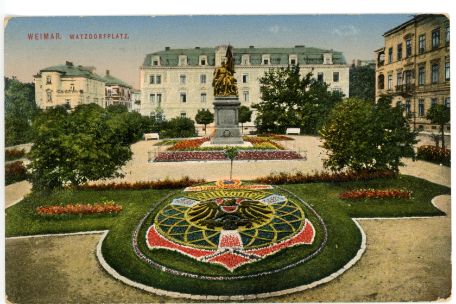 Es handelt sich um eine Postkartenmalerei. Zusehen ist ein Kriegsdenkmal auf der Mitte eines gestalteten Platz. Die gestelltung zeichnet sich durch aufwendig gepflegte Blumenbeete aus. Das Bild hebt die symetrische Gestaltung des Platzes nocheinmal hervor.