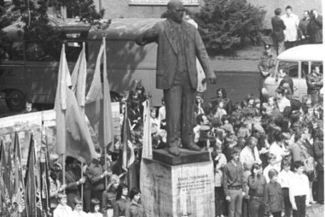 Im Zentrum des Bildes ist eine Statue von Ernst Thälmann zu erkennen. Um die Statue herum haben sich mehre Uniformierte Jugendliche mit Fahnen versammelt. Die Szene macht einen festlichen Eindruck.