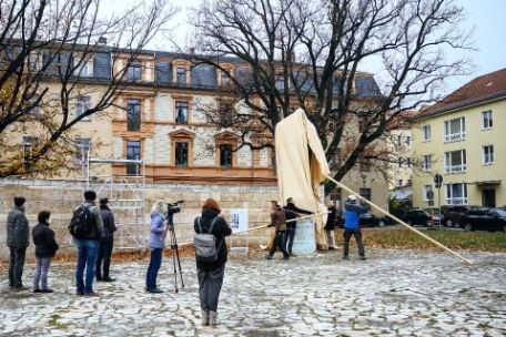 Zu sehen ist ein großerer Platz auf dem an die 10 Menschen stehen. Sie stehen verstreut auf dem Platz einer Verhüllten Statue zugewand. Einer der Zuschauer hat ein Stativ mit Kamera dabei welches Ebenfalls auf die verhüllte statue gerichtet ist. Unmittelbar an der Statue stehen Menschen die sich bereitmachen das Denkmal zu enthüllen.