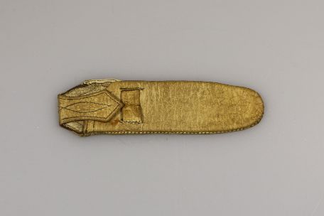 You can see a pocket knife case made from human skin. It has a yellowish colour due to the tanning process. It is rounded at both ends. On the right-hand side, you can see a folded and pointed clasp that has been folded onto the pouch.