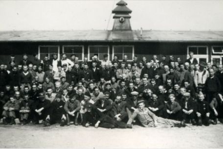 Group photo of liberated Dutch prisoners in front of a wooden barrack. Occasionally, some still wear prisoner clothing.