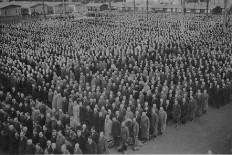 Auf dem überfüllten Buchenwalder Appellplatz stehen getrennt in achtziger Gruppen an die tausend Männer. Sie stehen in Reih und Glied in Blöcken von circa fünf mal 16 Personen, aber zwischen den Blöcken stehen noch weitere Menschen. Im Hintergrund einige Baracken.