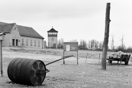 Im Vordergrund liegt auf der linken Seite eine Handplanierwalze. Rechts ist ein aufgerichteter Baumstamm zu sehen. Etwas hinter dem Pfahl steht ein Karre,n der mit Bruchsteinen beladen ist. Im Zentrum des Bildes steht ein Schild, welches dem Besucher informationen anbietet. Links im Hintergrund sind Eingang und Rechter Flügel der ehemaligen Häftlingskantine zu sehen. Rechts daneben steht einer der Wachtürme des ehemaligen Lagers.