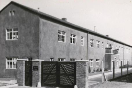 View of a two-story stone barracks, which is separately fenced. In the foreground of the picture, a wooden access gate can be seen between two stone pillars. To the left of it is a smaller one for people.