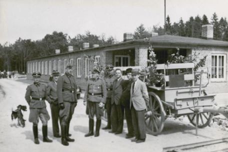 Lagerkommandant Pister (Mitte) und SS-Männer mit Betriebsleitern der Rüstungsfabrik. Die Männer stehen vor einem Pferdegespann. Im Hintergrund ein Bungalow.